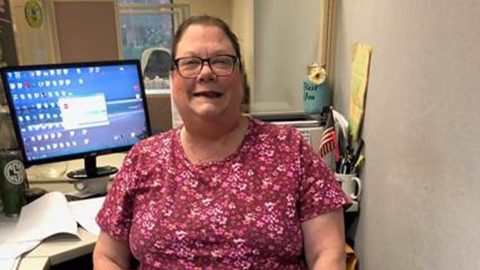 Carol, smiling, with her prosthetic at her computer desk.